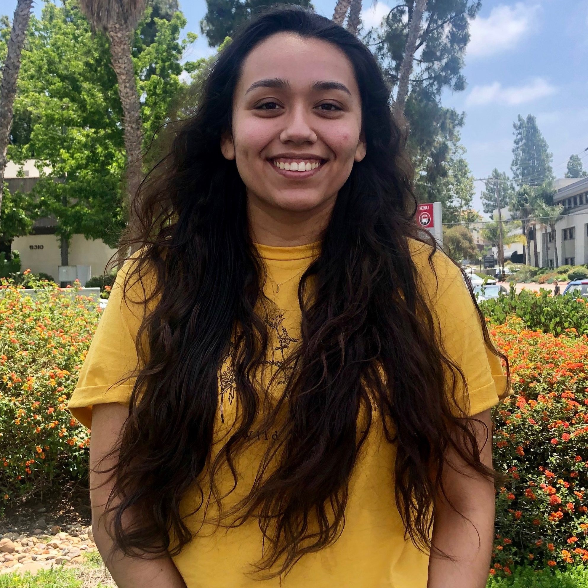 Headshot of Project Coordinator Brianna Hernandez wearing a yellow shirt and standing in front of flowers and trees