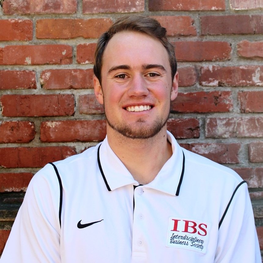 Headshot of former research assistant Isaac wearing a white shirt with a Nike check symbol on the right side of his shirt and the text reading the letters IBS in red meaning Interdisciplinary Business Society labeled underneath in black in front of a red brick wall background