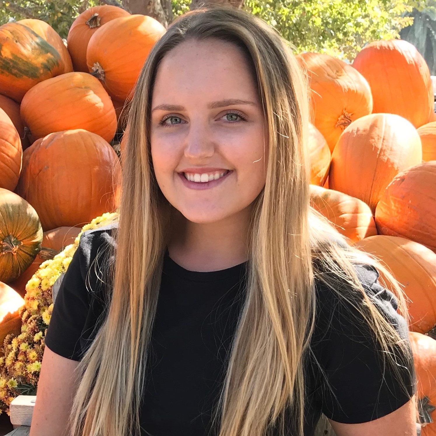 Headshot of former research assistant Katie S. with a background of sunlight shining on a hill of bright orange pumpkins