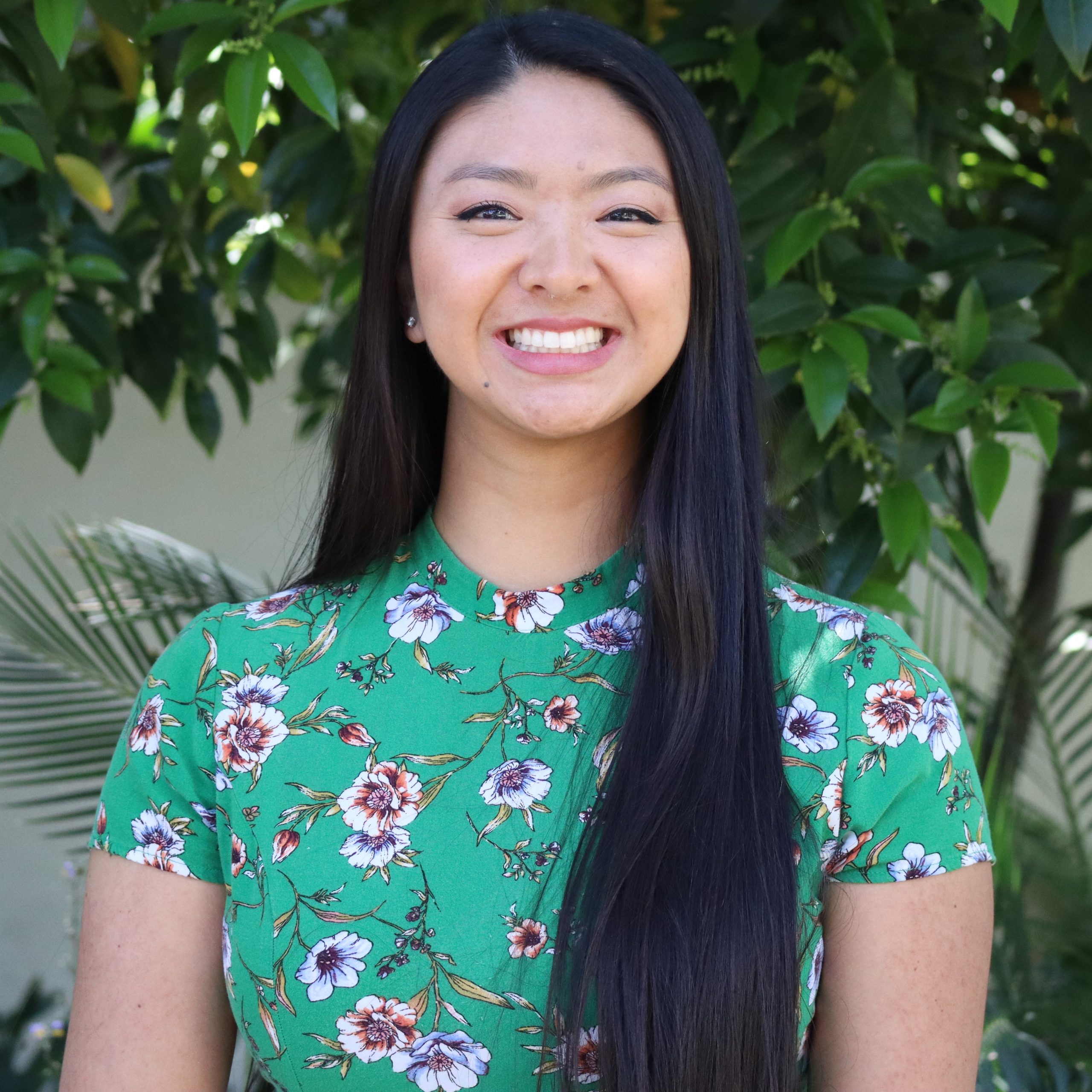 Headshot of research assistant Sabrena in a dark green floral dress in front of a green tree background