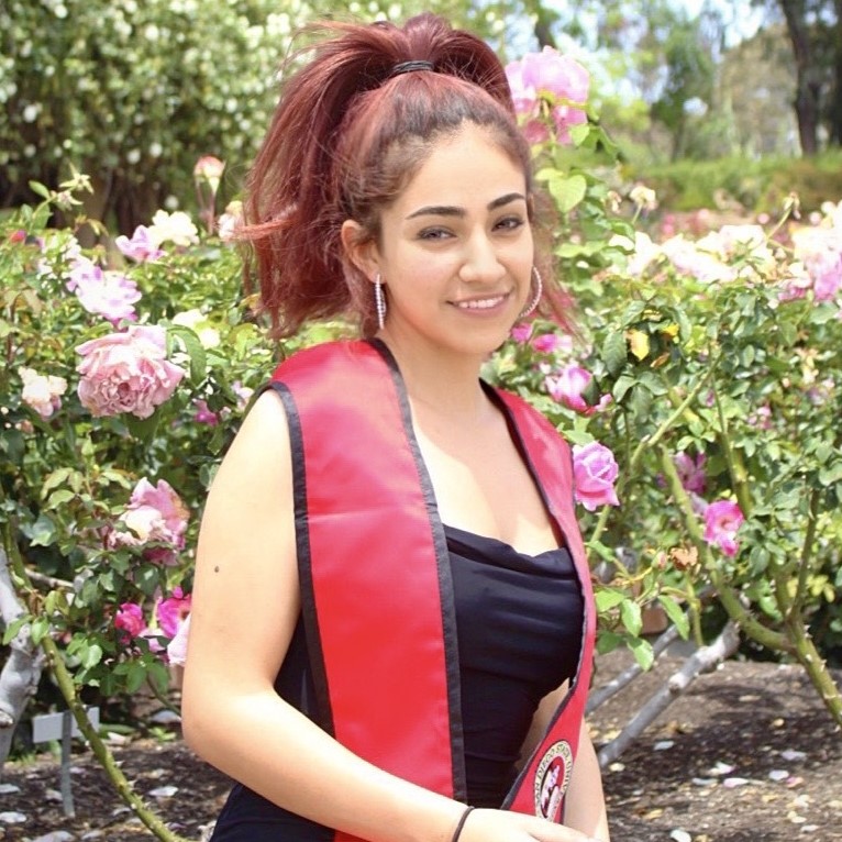 Headshot of research assistant Anna with dyed red hair, a black dress, and a red with black edge SDSU stole in front of green bushes with blossoming pink roses