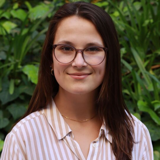 Headshot of Research Assistant Cassidy Loya wearing a brown and white striped blouse and glasses, standing in front of greet foliage