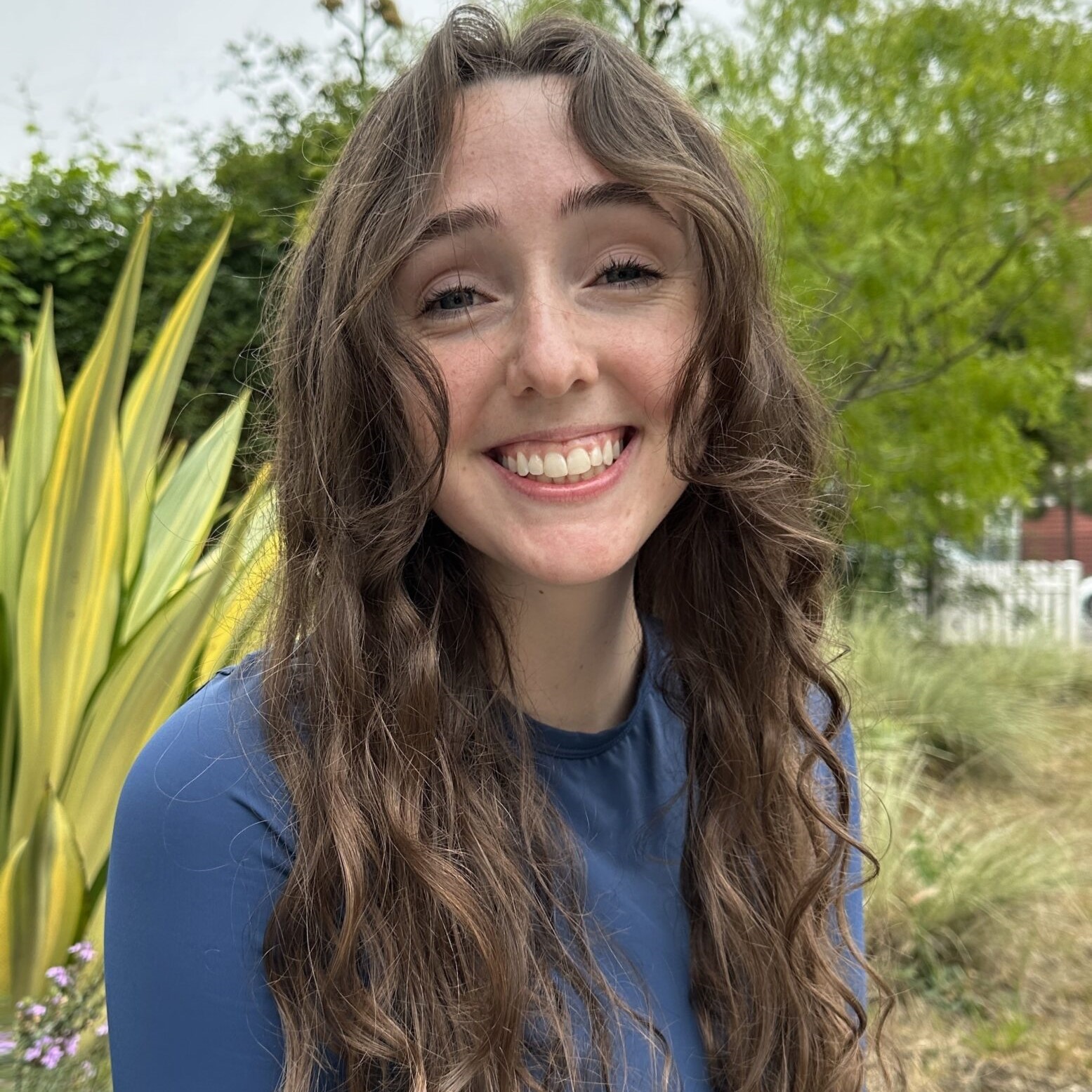 Headshot of Research Assistant Isabelle Magee wearing a blue shirt and standing in front of a green foliage