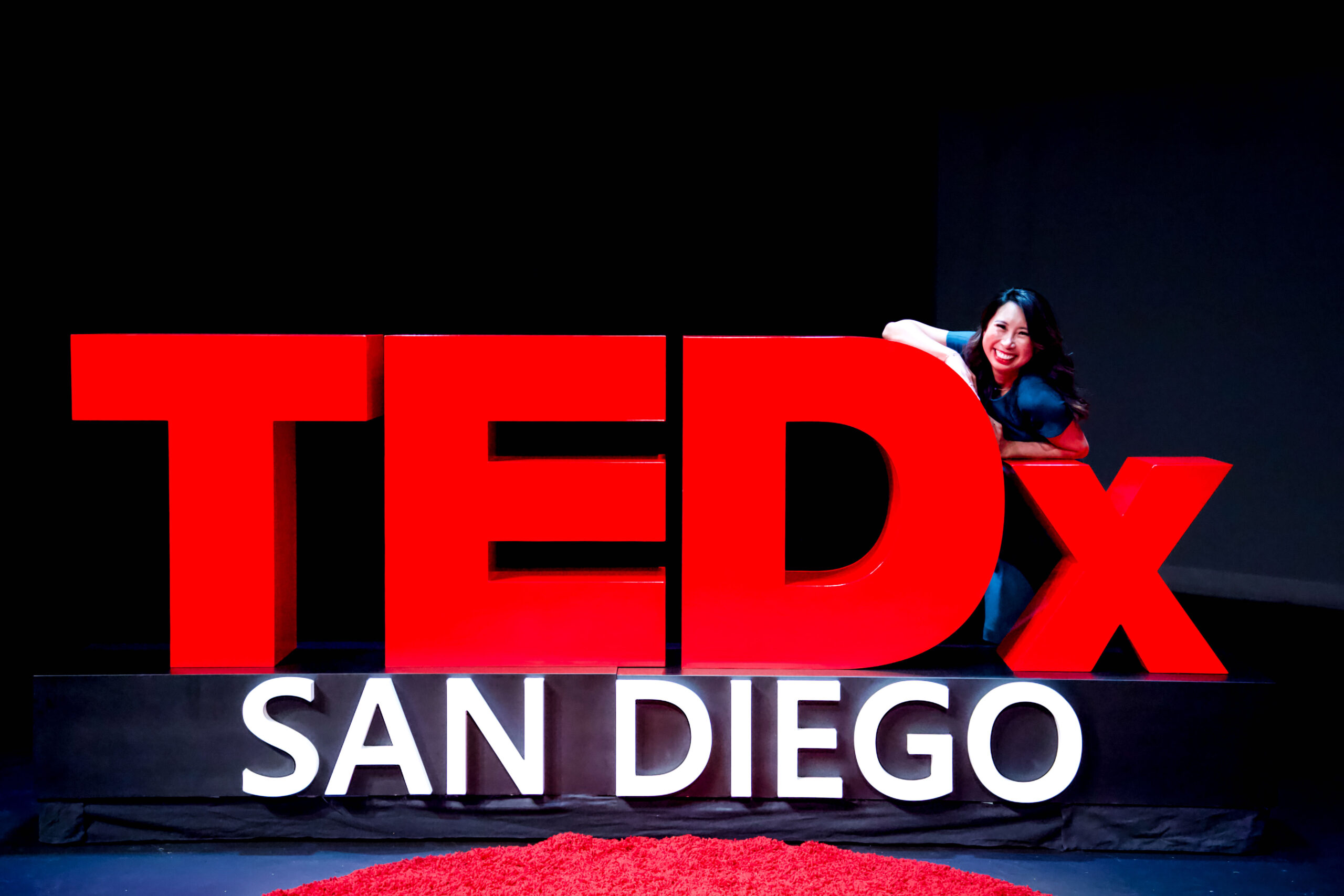 Image of Dr. Jillian Wiggins wearing a teal dress posing behind a TEDx sign.