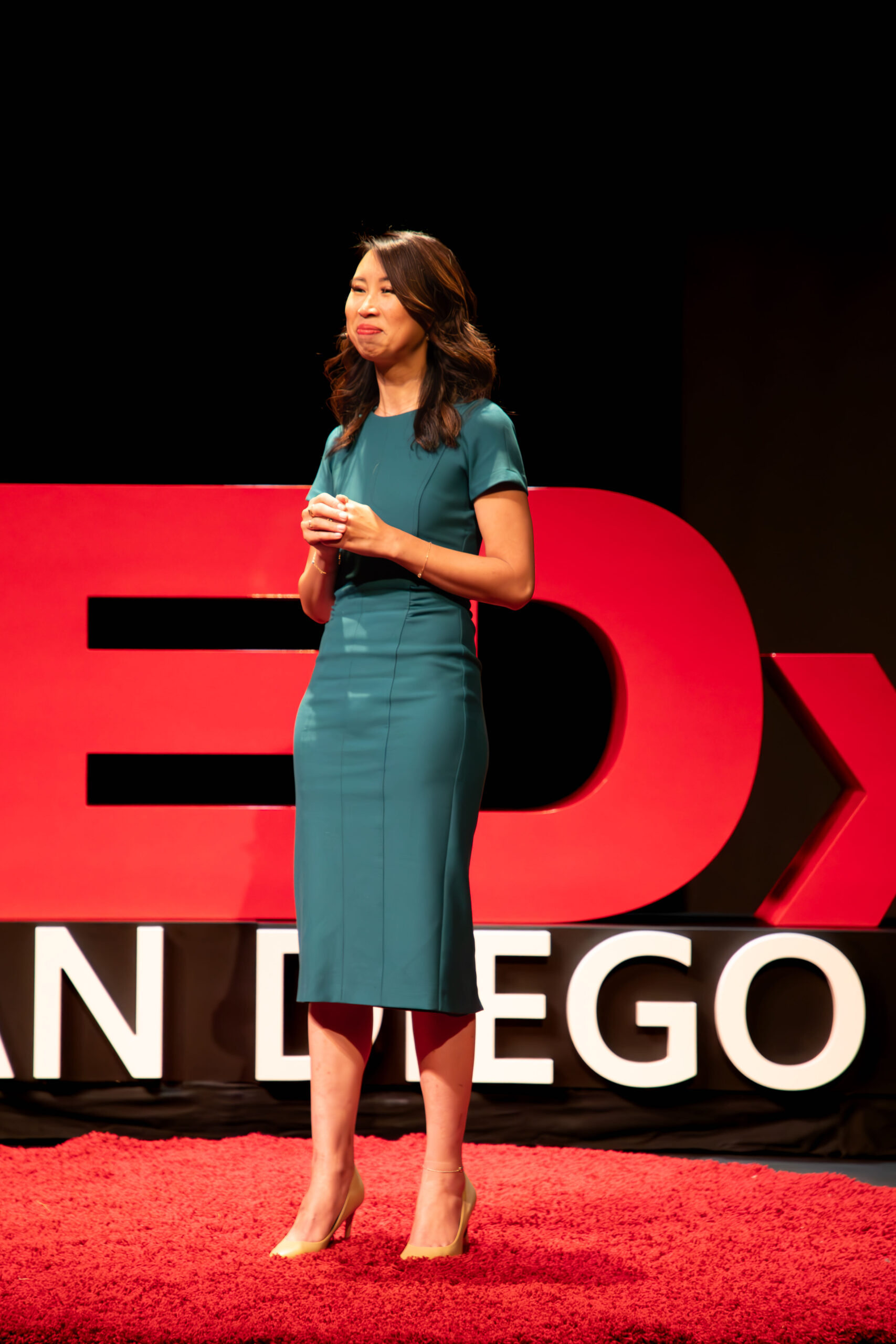 Image of Dr. Jillian Wiggins wearing a teal dress presenting in front of a TEDx background.