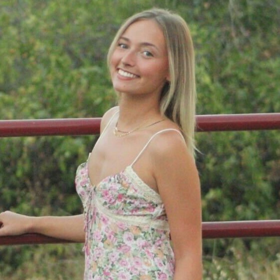 Headshot of research assistant Caitlin wearing floral dress smiling in front of red bar fence 