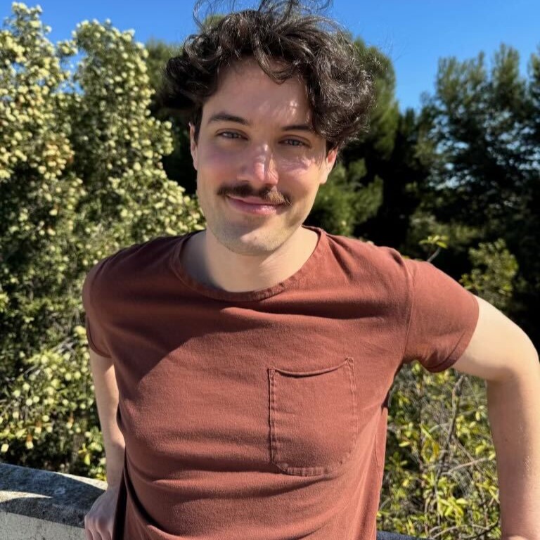 Headshot of research assistant Josh Syres wearing brick red t-shirt smiling outside in front of greenery