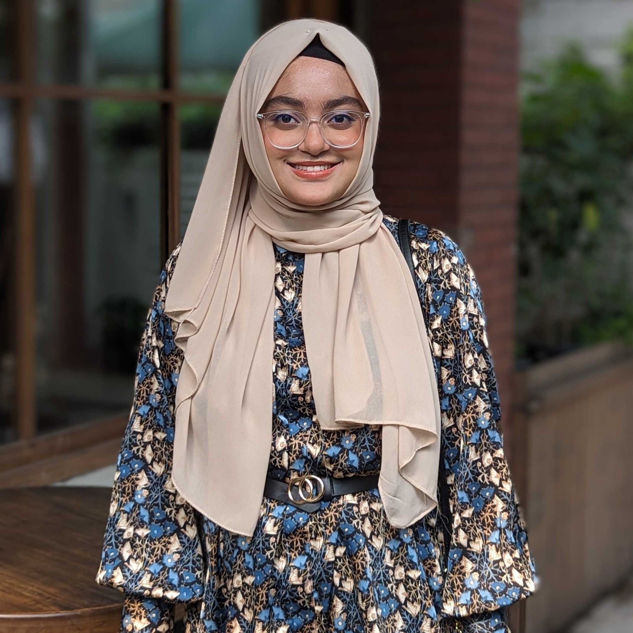 Headshot of master's student Munira wearing glasses in a floral long sleeve and beige headscarf, standing in front of a building