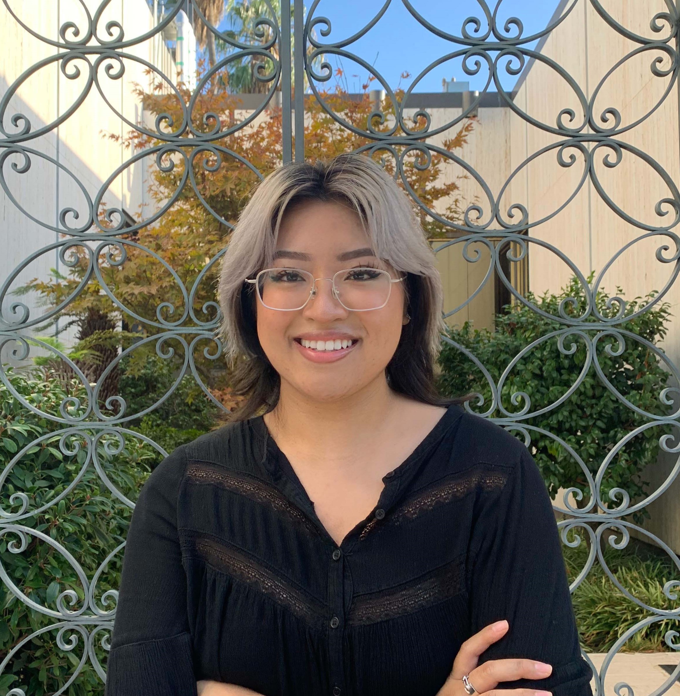 Headshot of Research Assistant Kayla Ta wearing a black shirt and standing in front of a gate & green foliage