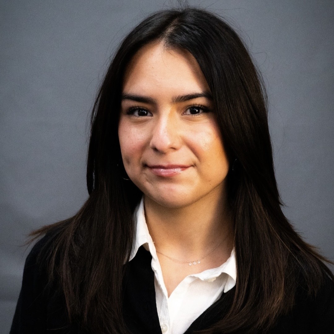 Headshot of research assistant, Rose Gomez, in a white button-up shirt with a black jacket with hair down, standing and smiling indoors