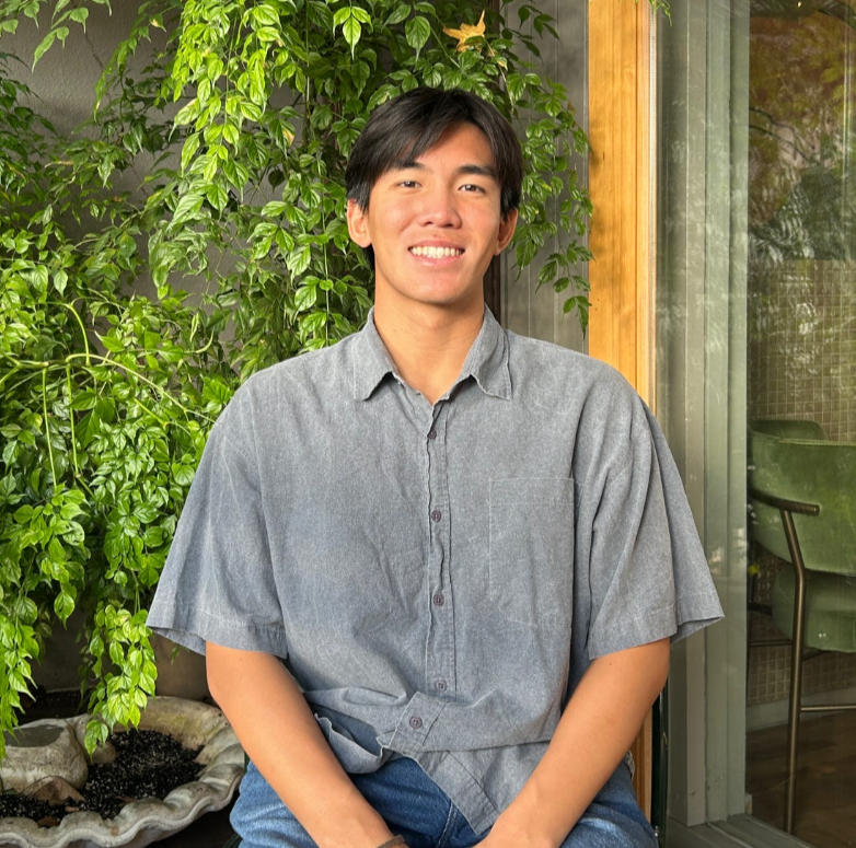 Logan Phan sitting on a chair, posing for a photo in front of a background of green plants